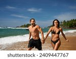 Fit couple runs on tropical beach, playful man and woman exercise near ocean. Athletic pair in swimwear enjoys summer vacation, jogging along shoreline, healthy lifestyle, beach workout, sunny day.