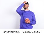 Small photo of Upset depressed young arab man with curly hair wearing purple sweatshirt over white background makes face palm as forgot about something important holds mobile phone expresses sorrow and regret blames