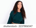 Small photo of Beautiful teen girl with curly hair wearing green sweater over white background with snobbish expression curving lips and raising eyebrows, looking with doubtful and skeptical expression, suspect.