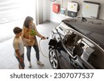 Small photo of Happy son and mother plug EV charger from home charging station to electric vehicle. Future eco-friendly car powered by renewable source of clean energy on daytime. Horizontal, high angle.