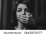 Portrait of a young suffering sad woman with tears in her eyes, a man closes her mouth with his hand. Domestic violence, crying, religion, disagreement, fight, divorce, beating a weaker person, dark.