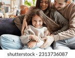 Portrait of happy smiling adorable little daughter in gentle embrace of young loving parents enjoying family lifestyle games at cozy home living room. Cheerful husband keeping wife and child in arms
