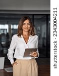 Small photo of Vertical portrait of Latin mature adult professional business woman looking at camera and smiling. European businesswoman CEO holding digital tablet using fintech tab application standing in office.