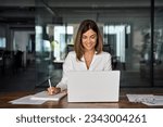 40s mid age European business woman CEO working on laptop sitting at table workspace, making notes in office. Smiling Latin Hispanic mature adult professional businesswoman using pc digital computer