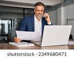 Smiling Hispanic businessman talking phone and check database in office. Happy Latin or Indian male business man holding documents, working at laptop computer doing online trade market tech research.