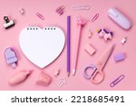 White heart shaped notebook on a pink background surrounded by stationery, markers, erasers, pencils, paper clips, pens, scissors. Back to school concept, splash screen for online stationery store.