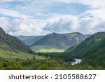 Small photo of Colorful view to sunlit green mountain valley with forest and river against mountain range under cloudy sky. Wide mountain valley in sunlight and large mountains in lush clouds in changeable weather.