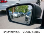The left side black mirror of a passenger car, which reflects the cars behind standing in two rows on the road during the day.