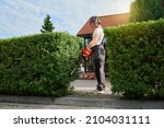 Competent caucasian gardener cutting bushes with electric trimming machine on back yard. Strong young man using modern gardening tool for shaping overgrown hedge.