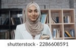 Female muslim student in library, holding books and looking at camera, girl wearing hijab spending time studying - modern Islam, student lifestyle concept close up 