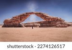 Small photo of A "Dust Devil" or small tornado passes through "Rainbow Rock", also known as "Arch Rock' in Alula, Saudi Arabia. A man wearing traditional Clothes stands right under the arch.