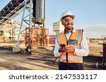 Small photo of A young African mine worker wearing protective wear is looking off camera while holding a cell phone with coal mine equipment in the background
