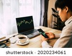 Small photo of Asian young man 30s looking stock tickers or graphs cryptocurrency trading platforms on smartphone and laptop with a cup of coffee, calculator, notebook on table at home office in the morning.