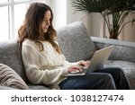 Smiling attractive young woman sitting on sofa using laptop communicating working online at home, happy teen girl typing on computer, enjoying writing blog or chatting with friends in social network