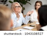 Attractive aged businesswoman, teacher or mentor coach speaking to young people, senior woman in glasses teaching audience at training seminar, female business leader speaker talking at meeting