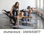 Young female yoga instructor teaching Bird dog pose, Knee to Forehead curl exercise for a group of sporty people practicing in studio, working out indoor, teacher helping to master, full length