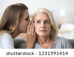 Shocked older woman listening to young female whispering in ear, friend gossiper telling secret rumors or unbelievable news to surprised senior lady, old mother and adult daughter gossiping concept