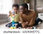 Happy cute little son playing game with black dad baby sitter building constructor tower from multicolored wooden blocks, african family father and toddler child boy having fun on warm floor at home