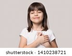 Little sincere adorable girl closed eyes holding hands on chest feeling gratitude pose isolated on sandy color beige background, arms on heart gesture of love appreciation gratitude, adoption concept