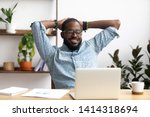Smiling Afro-American businessman holding hands behind head sitting at office desk behind laptop. Happy black employee feeling no stress, relaxing, watching funny video after successful working