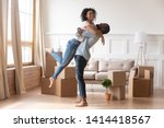 Happy african american couple first time home buyers celebrate new house purchase on moving day, smiling husband embracing lifting excited young wife laughing standing among boxes in own flat house