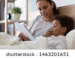Mixed race african american family mom baby sitter and cute little kid son read book fairy tale in bed, cute small adopted child boy listening to mom foster parent learn reading in bedroom in morning