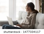 Small photo of Thoughtful young woman in eyeglasses sitting with computer on couch, looking outside, cannot concentrate on work, need some rest, feeling bored, need additional motivation, working remotely at home.