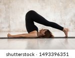 Young woman wearing black sportswear practicing yoga, doing Halasana exercise, standing in Plough pose, beautiful sporty girl with closed eyes working out at home or in yoga studio with grey walls