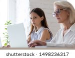 Small photo of Surprised young woman worker sit at office desk work near coworker feel stunned amazed looking at senior colleague laptop screen, shocked female employee spy peep at workmate computer in office