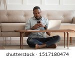 African guy sits leaned on couch cross-legged on carpet near coffee table hold cellphone distracted from work chat online, self-employed man solve business issues contact with client distantly concept