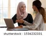 Happy asian muslim businesswoman sales manager shake hand of caucasian lady client make deal with female customer at meeting with laptop, diverse women partnership, respect and collaboration concept