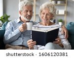 Small photo of 65s couple enjoy time with literature and favourite beverage, wife read book while husband hug her and drink coffee, spouses booklovers having common interests and hobby, like-minded family concept