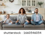 Calm young family with little daughter sit on couch practice yoga together, happy parents with small preschooler girl child rest on sofa meditate relieve negative emotions on weekend at home