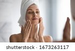 Small photo of Closeup head shot pleasant beautiful woman applying moisturizing creme on face after shower. Smiling young pretty lady wrapped in towel smoothing perfecting skin, daily morning routine concept.