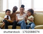 Happy african american family and children watching funny tv show or movie eating popcorn snack. Happy diverse dad holding remote controller, mom hugging cute siblings.