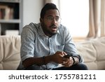 Small photo of African millennial guy makes big eyes amazed shocked face expressions sitting on couch spending free time holding remote control watching thriller scary movie feels appalled, unbelievable news concept
