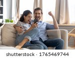 Overjoyed surprised young couple looking at phone screen, reading message with good news, excited woman and man celebrating success, online lottery win, showing yes gesture, sitting on couch