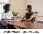 Smiling African American young businesswoman talk negotiate with female Caucasian job candidate at office meeting, happy biracial boss discuss ideas with business partner or employee at interview