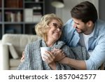 Smiling mature woman holding cup, hugging with adult son close up, enjoying leisure time, happy elderly mother wearing warm scarf drinking hot tea or coffee, family spending weekend together at home