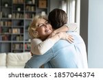 Close up smiling mature woman wearing glasses hugging adult son, standing in living room at home, family enjoying tender moment, beautiful happy middle aged woman embracing young man