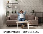 Peaceful young man relaxing meditating on comfortable sofa with folded hands behind hand, breathing fresh air, daydreaming napping enjoying calm mindful lazy weekend moment alone in modern living room