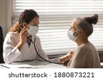 Small photo of Caring young female doctor in medical facemask hold stethoscope listen to elderly patient heart in hospital. Woman GP or physician do regular checkup examine mature client at consultation in clinic.