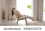 Small photo of Happy millennial Caucasian woman sit in cozy chair in design home stretch relax breathe fresh air. Smiling young female renter or tenant rest in armchair relieve negative emotions enjoying weekend.