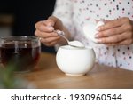 Just one teaspoon is enough. Close up cropped shot of young lady brew strong black herbal flower tea add heaped spoon of granulated sugar for a cup. Focus on transparent teacup with hot drink and bowl