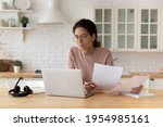 Thoughtful hispanic female sit at home workplace hold financial documents look on pc screen check data. Serious young woman think on business offer compare terms conditions in paper letters and online