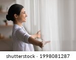 Overjoyed young Indian woman open curtains look in window distance meet welcome new day at home. Smiling millennial mixed race female feel excited about life career opportunities or perspectives.