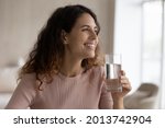 Smiling young Hispanic woman enjoy clean mineral water from glass for body refreshment. Happy Latino female drink clear pure mineral still water follow healthy lifestyle. Diet, habit concept.