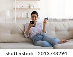 Excited Indian woman paying online by credit card for goods, using smartphone, sitting on couch at home, happy young female satisfied customer browsing internet banking service on device, shopping