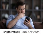 Doubtful young man sit on sofa hold phone look aside hesitate in making important choice decision before make call. Worried concerned millennial businessman think on disturbing news received by email