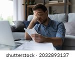 Frustrated desperate millennial man checking bills for payments, holding receipts, getting upset about overspending, too high mortgage, insurance fees. Homeowner analyzing costs, expenses, budget